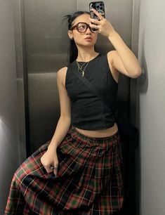 a woman taking a selfie with her cell phone in an elevator stall wearing a plaid skirt and black crop top