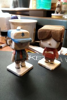 two wooden toy figurines sitting on top of a table next to each other