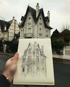 a person holding up a drawing in front of a large white house with black roof