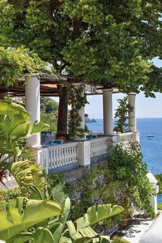 an outdoor dining area overlooks the ocean and is surrounded by greenery, with white pillars