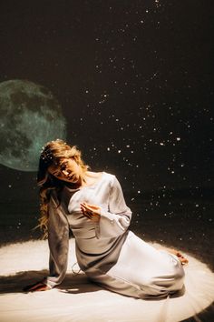 a woman sitting on the ground in front of a full moon and stars filled sky