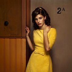 a woman in a yellow dress leaning against a wall with her hand on her shoulder