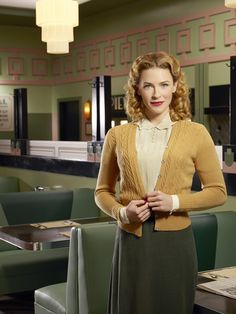 a woman standing in front of a table wearing a yellow sweater and green skirt with her hands on her hips