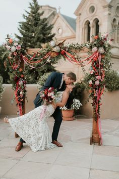 a bride and groom kissing under an arch decorated with red ribbon, flowers and greenery