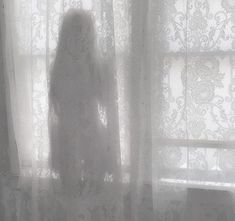 a woman standing in front of a window next to a sheer curtain with white lace on it