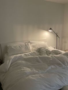 an unmade bed with white sheets and pillows in a small room next to a lamp