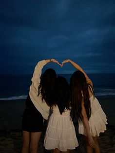 two girls standing on the beach making a heart shape with their hands while they both look at the ocean