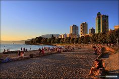 many people are sitting on the beach by the water and buildings in the background at sunset