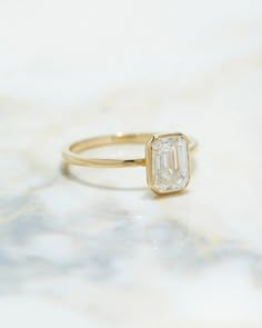 a gold ring with an emerald cut diamond on the front, sitting on a marble surface