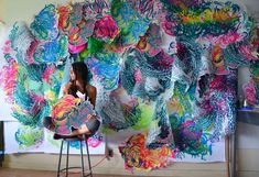 a woman sitting on a stool in front of a wall covered with colorful paper flowers