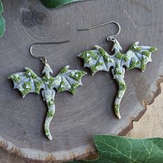 two dragon shaped earrings on top of a tree stump with green leaves around them and one is made out of polymer