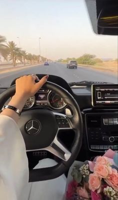 a woman driving a car on the road with flowers in front of her and palm trees behind her