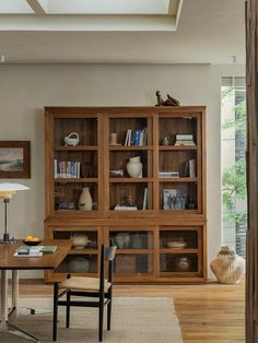 a wooden bookcase with glass doors in a living room next to a table and chair