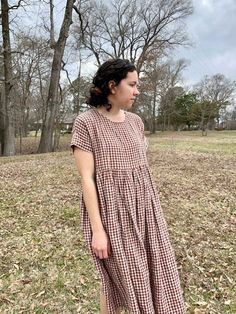 a woman standing in the grass wearing a brown and white checkered dress with short sleeves