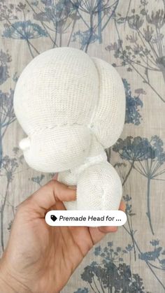 a hand holding a white knitted teddy bear in front of a floral wallpaper