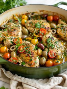 a pan filled with chicken, olives and tomatoes on top of a table next to a cloth