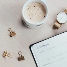 a cup of coffee next to a notebook and some gold clips on a table with a watch