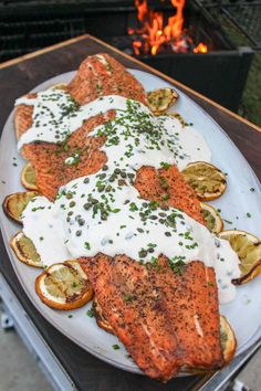 grilled salmon with white sauce and lemons on a platter next to an open fire