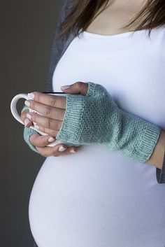 a pregnant woman holding a coffee cup in her hand while wearing knitted mitts