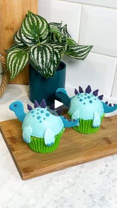 two dinosaur cupcakes sitting on top of a cutting board next to a potted plant