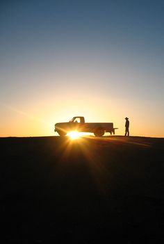 a man standing next to a truck on top of a grass covered field at sunset