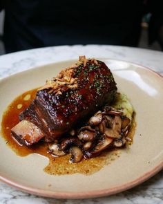 a piece of meat with mushrooms and sauce on a white plate sitting on a marble table