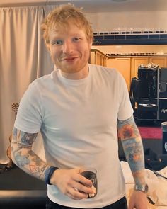 a man with red hair and tattoos holding a cup in his hand while standing next to a bed