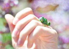 a small green frog sitting on top of a person's hand in front of purple flowers