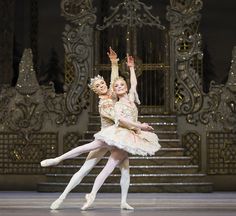 two ballerinas in white tutus and gold tiaras, one holding the other's leg