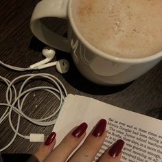 a woman's hand with red nail polish next to an open book and headphones