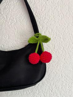 a crocheted cherries hanging from a black purse on a white wall background