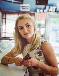 a woman sitting at a table with a piece of food in her hand and looking into the camera