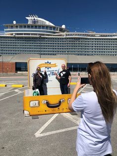 a woman taking a photo of two men in front of a large cruise ship with a yellow suitcase