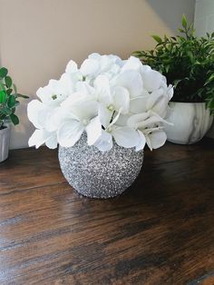 some white flowers are sitting in a silver vase on a wooden table next to potted plants