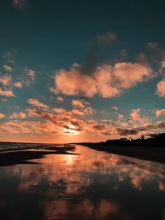 the sun is setting at the beach with kites flying in the sky over the water