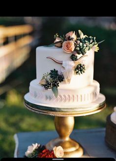 a white wedding cake with flowers on the top is sitting on a gold stand outside