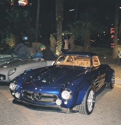 two classic cars parked next to each other on a sidewalk at night time with people sitting in the background