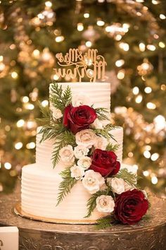 a white wedding cake with red and white flowers on top, sitting in front of a christmas tree