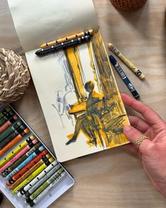 a person holding a pencil next to some crayons and a drawing on a piece of paper