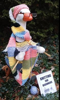 a knitted duck sitting on the ground next to a sign that says knitting for beginners