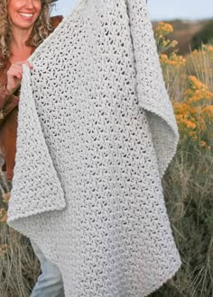 a woman holding up a white crocheted blanket in the desert with wildflowers behind her