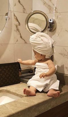 a baby sitting on top of a bathroom counter next to a sink with a towel wrapped around it's head