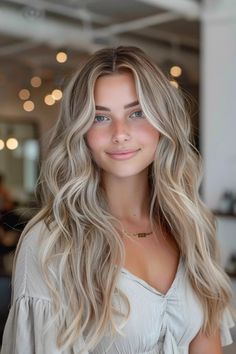 Woman with long blonde wavy hair and a necklace smiling indoors with blurred lights in the background. Hombre Blonde Hair, Soft Summer Balayage, Blonde Hair Inspo Pics, Coconut Cream Blonde Hair, Blonde Hair Length Ideas, Fall Hair Inspo For Blondes, Blonde For Pale Skin Blue Eyes, Natural Blonde Hair Inspiration, Blonde And Dark Highlights