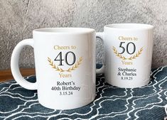 two white coffee mugs sitting on top of a blue and white table cloth with the words cheers to 40 years printed on them