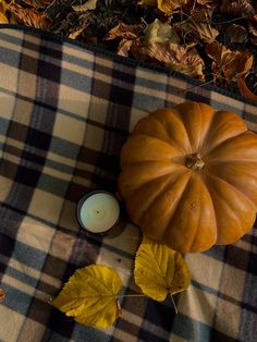 a candle and pumpkin sitting on a plaid blanket next to leaves in the fall season