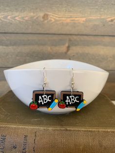 a pair of earrings with the words abc and apple on them sitting in front of a bowl