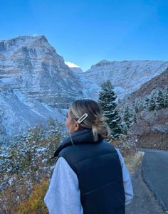 a woman standing on the side of a road looking at snow covered mountains and trees