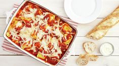 a casserole dish with meat, cheese and sauce next to bread on a table