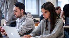 two people working on their laptops in an office setting with other people sitting at tables