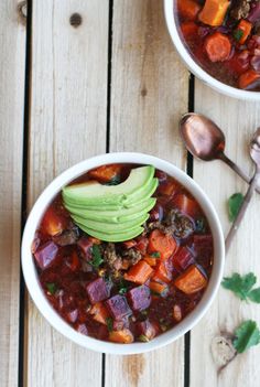 two bowls filled with chili and vegetables on top of a wooden table next to spoons